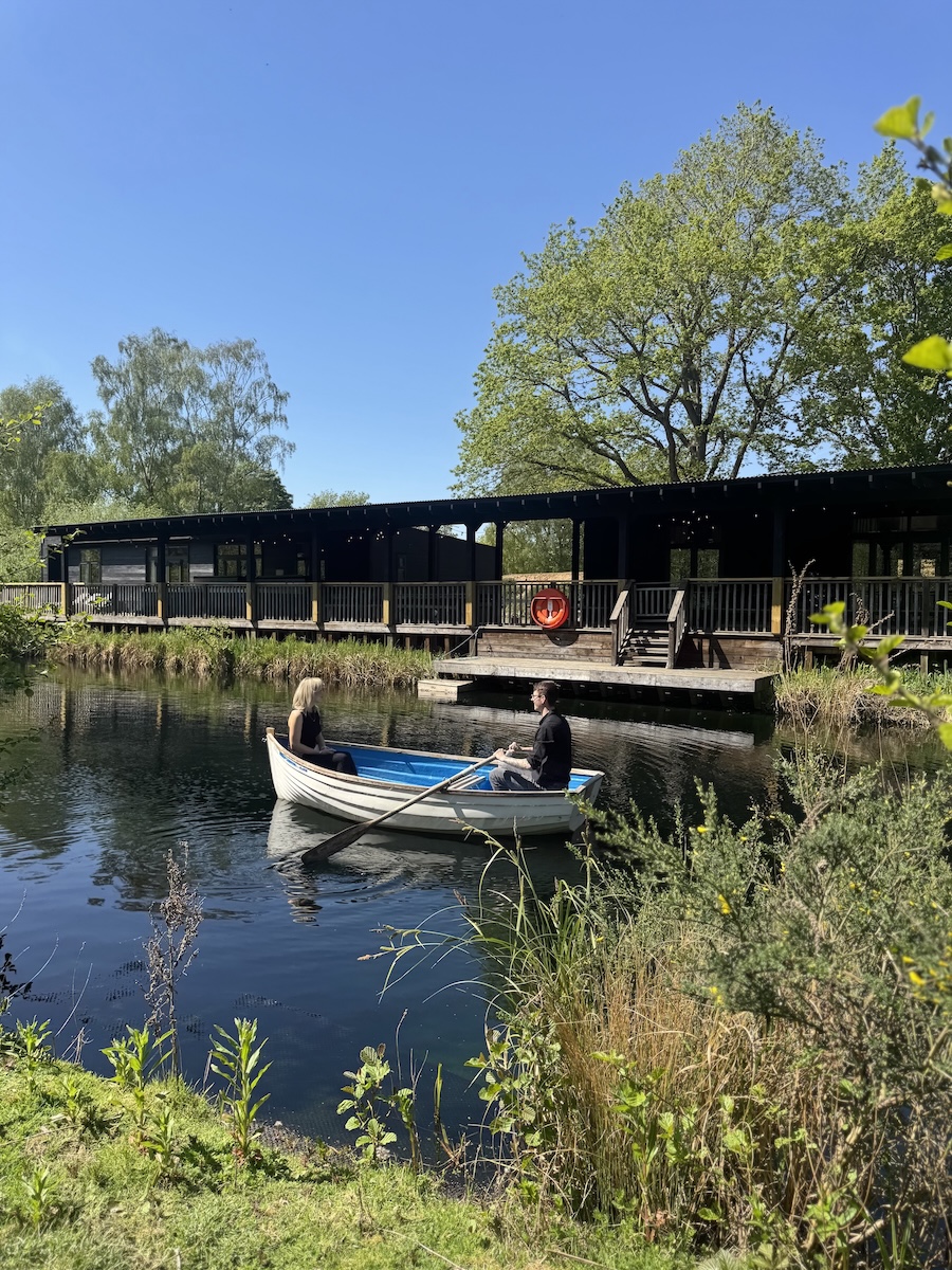 Two people row a small white and blue boat on a calm canal beside a wooden riverside building with a lifebuoy on the railing under a clear blue sky in daylight.