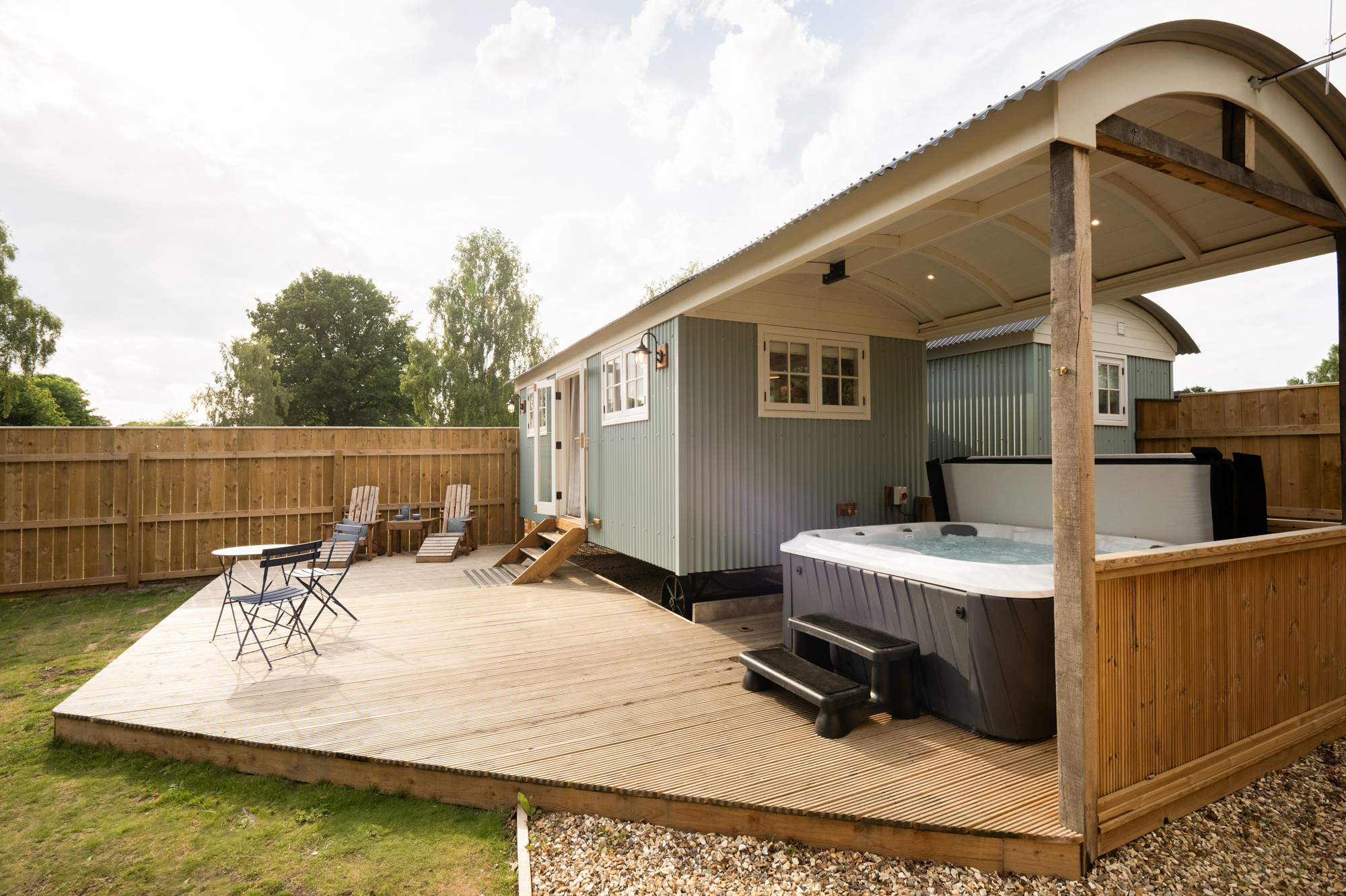 Backyard deck with a blue-green shed, hot tub, and wooden fencing; several chairs and a table sit on the deck with trees in the background.