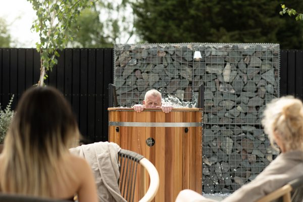 Man soaking in a wooden tub while water splashes, audience in foreground during an outdoor demonstration.