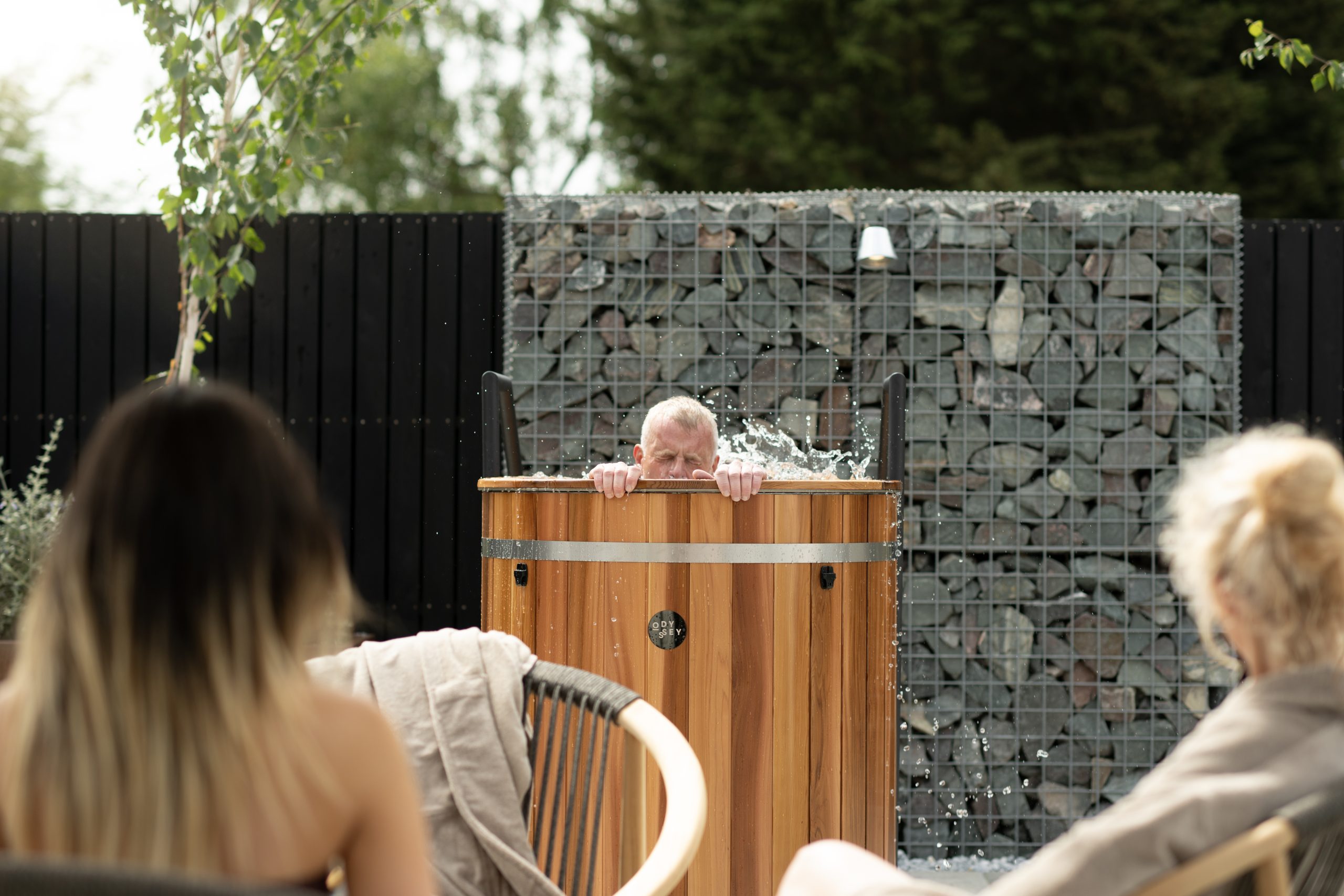 Man soaking in a wooden tub while water splashes, audience in foreground during an outdoor demonstration.
