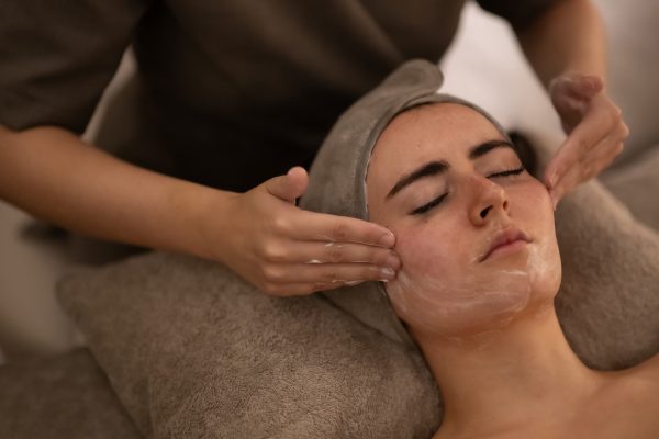 Person lying with eyes closed as a therapist gives a facial massage, hands on cheeks and a towel wrapped around their head.
