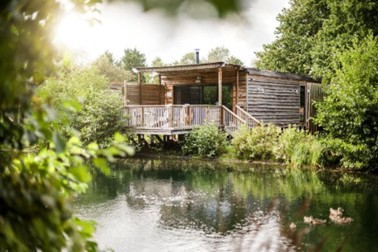 Wooden cabin on stilts over a calm pond, surrounded by trees and greenery with sunlight filtering through foliage.