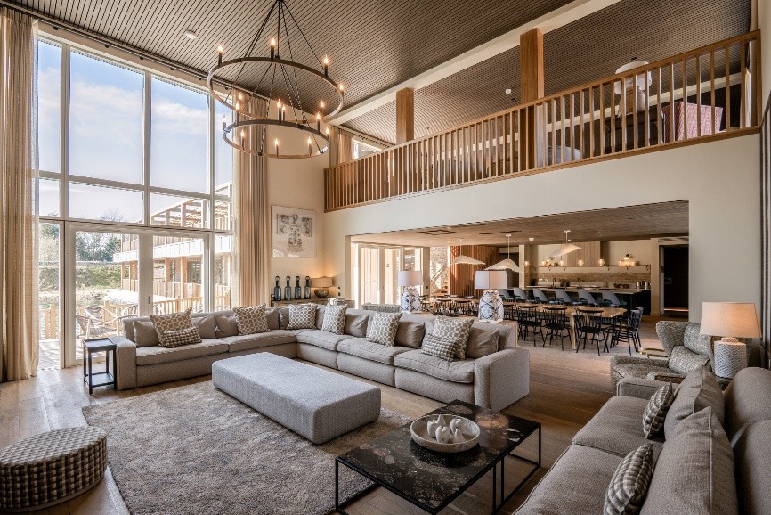 Spacious open-concept living room with a large gray sectional, coffee table, and floor-to-ceiling windows overlooking a dining area and mezzanine loft.
