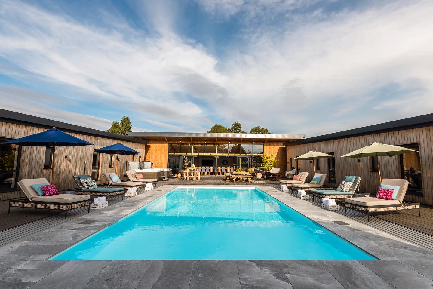 Resort-style pool area with loungers, umbrellas, and a modern wooden building in the background under a blue sky.