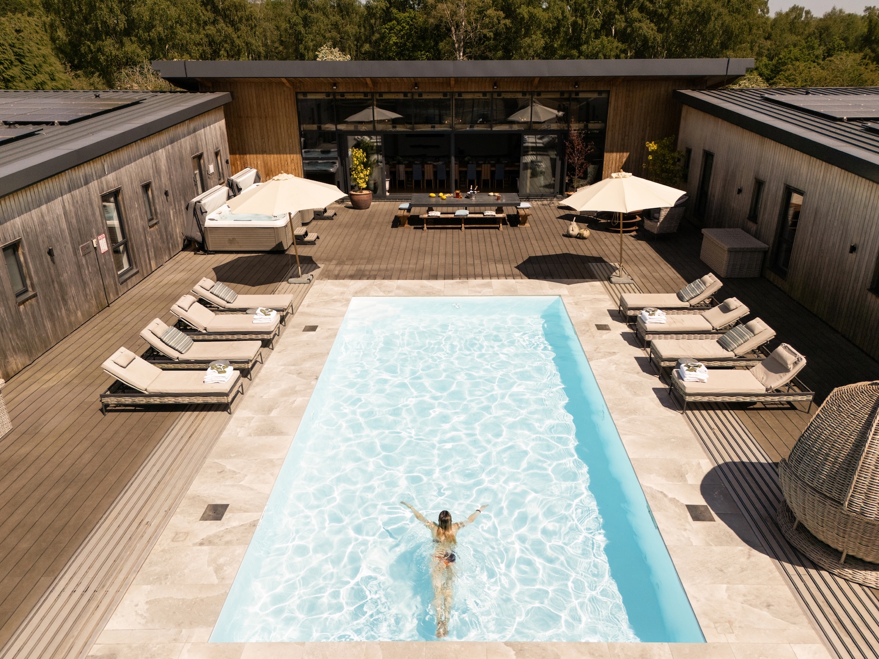 Rectangular outdoor pool surrounded by lounge chairs and umbrellas on a wooden deck, with a modern building backdrop and a person swimming mid-pool.