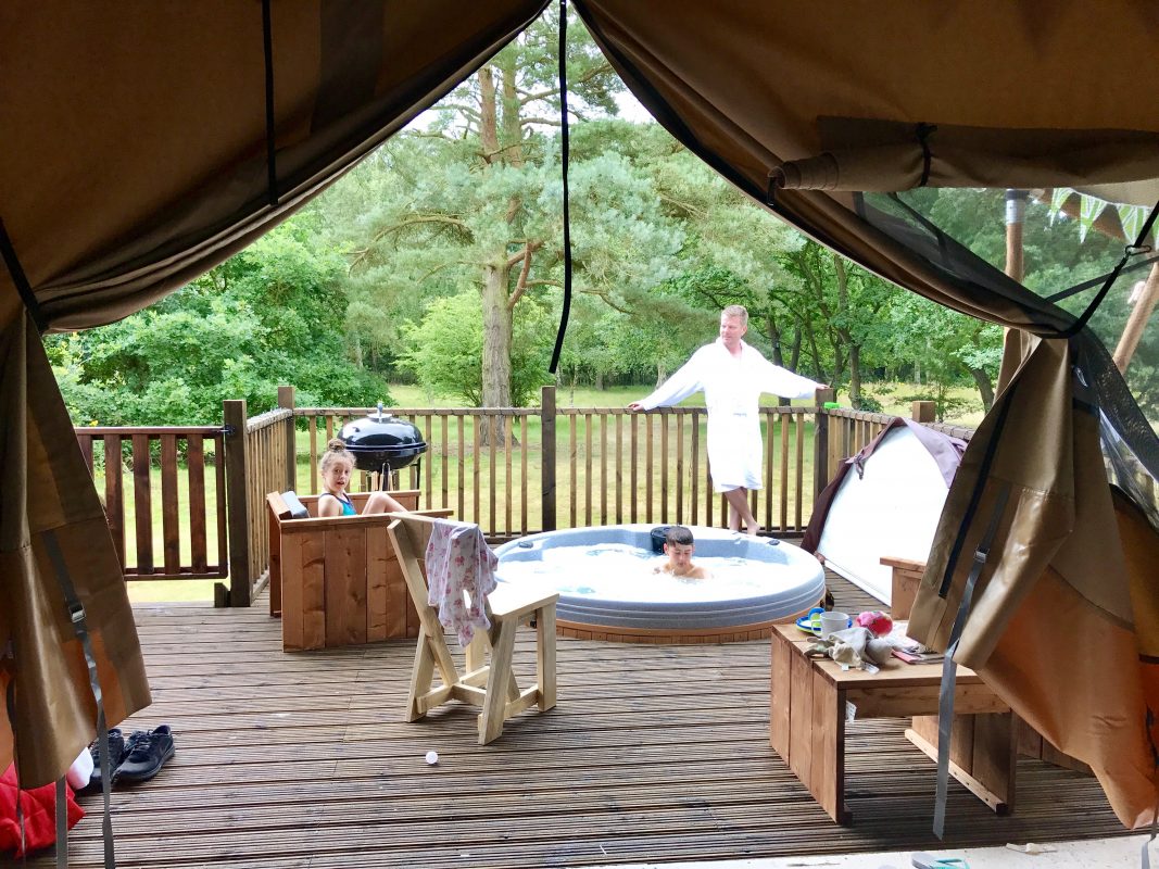 Family on a wooden deck with a round hot tub; two kids nearby and a man in a white robe stands at the railing, camping gear in view.