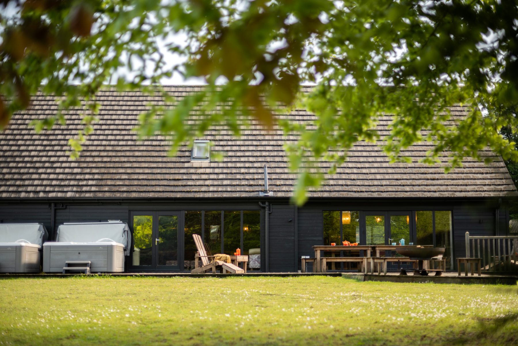 Backyard view of a modern house with a dark wooden exterior, large glass doors, and a patio table under a leafy canopy.