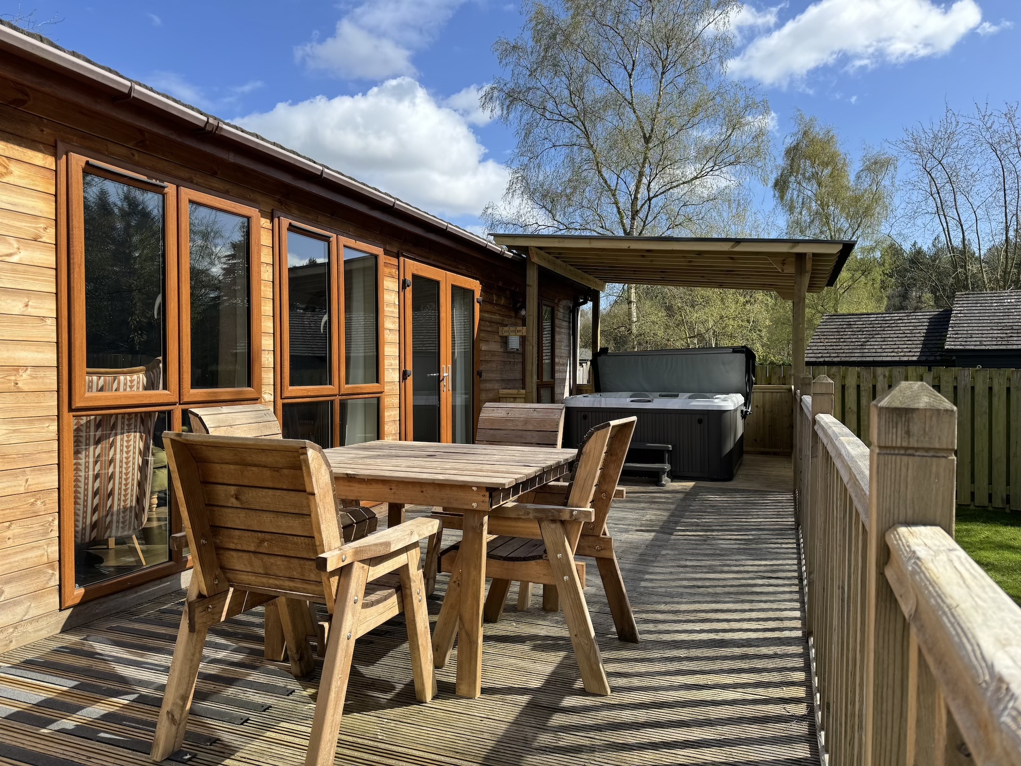 Wooden deck with outdoor dining set beside a wood-clad house, under a blue sky with trees in the background.