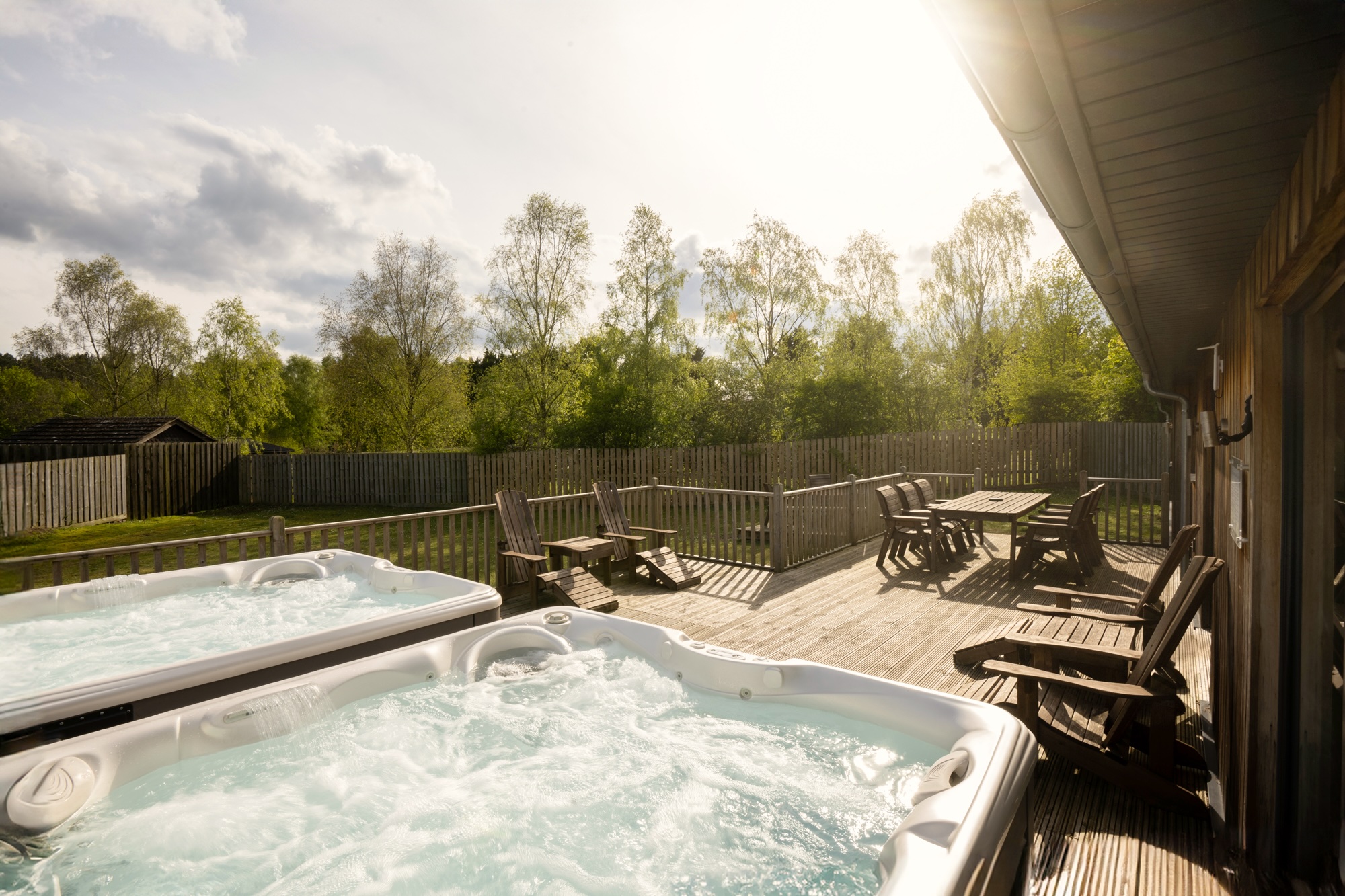 Wooden deck patio with two hot tubs, surrounding wooden chairs, a table, and a fenced yard with trees in sunlight.