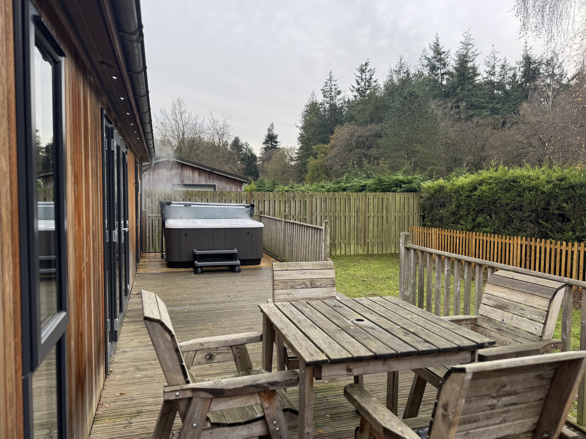 Wooden deck attached to a house with a hot tub and picnic-style tables, overlooking a fenced yard and treeline at a cloudy day.