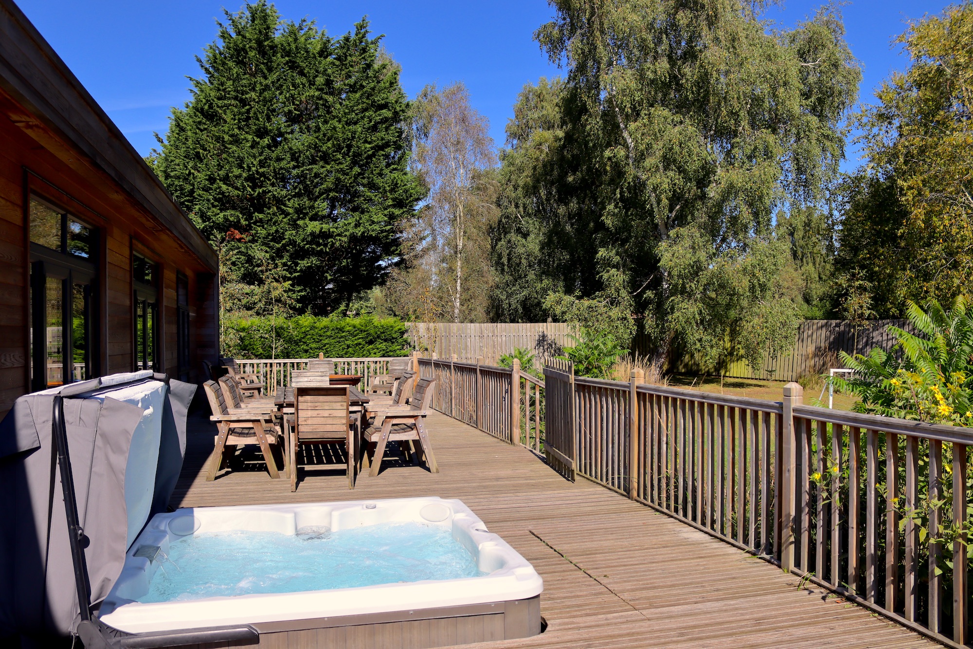 Wooden backyard deck with outdoor dining table and chairs beside a covered hot tub; fence and tall trees under a blue sky.