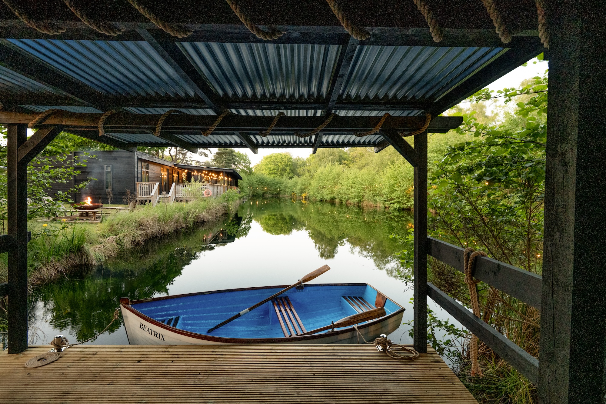 Blue and white boat named Beatrix moored under a wooden dock over calm water, with a cabin and string lights along the bank in the background.