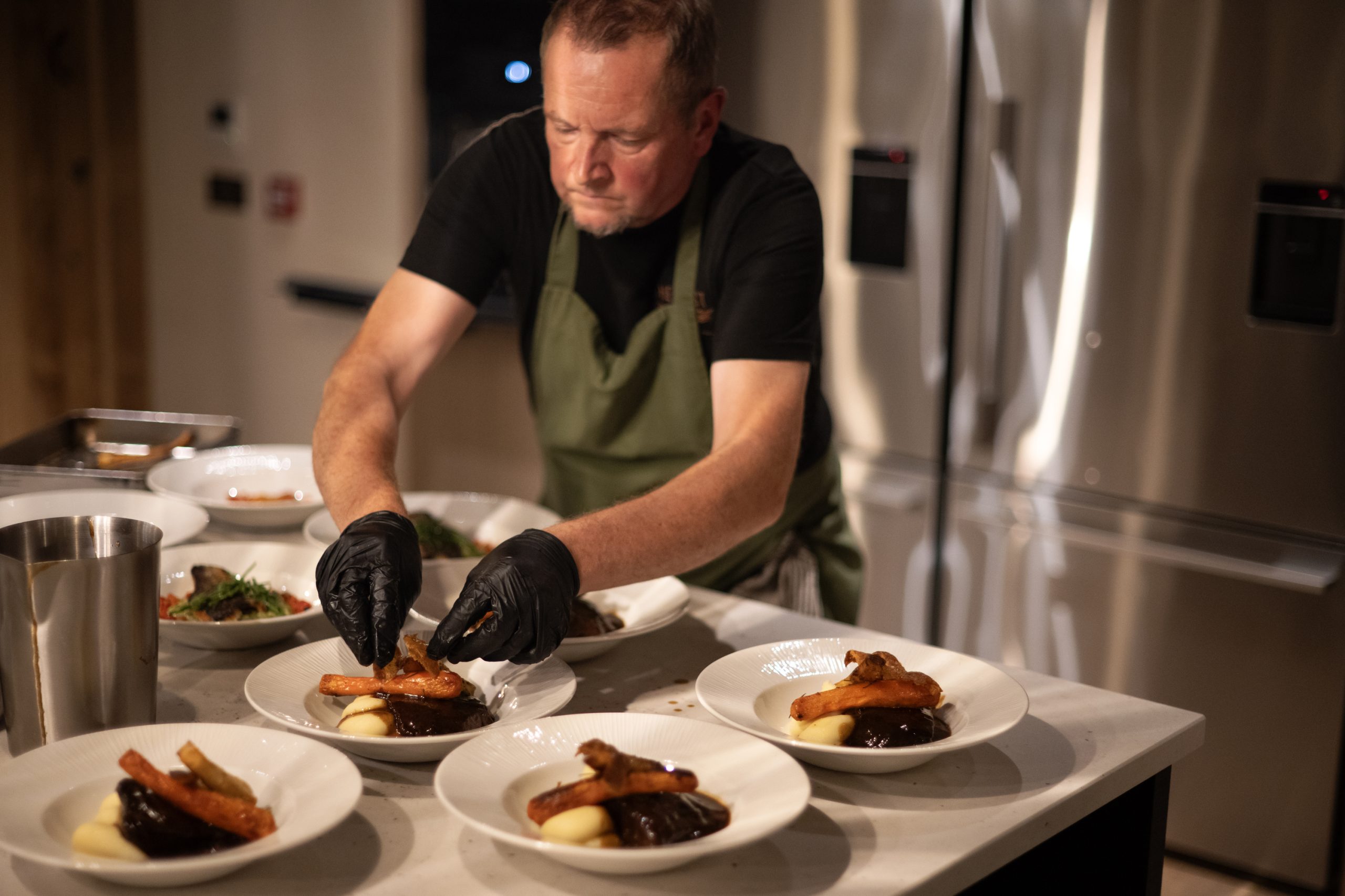 Chef in green apron and black shirt plating dishes at a stainless-steel counter in a modern kitchen, wearing black gloves.